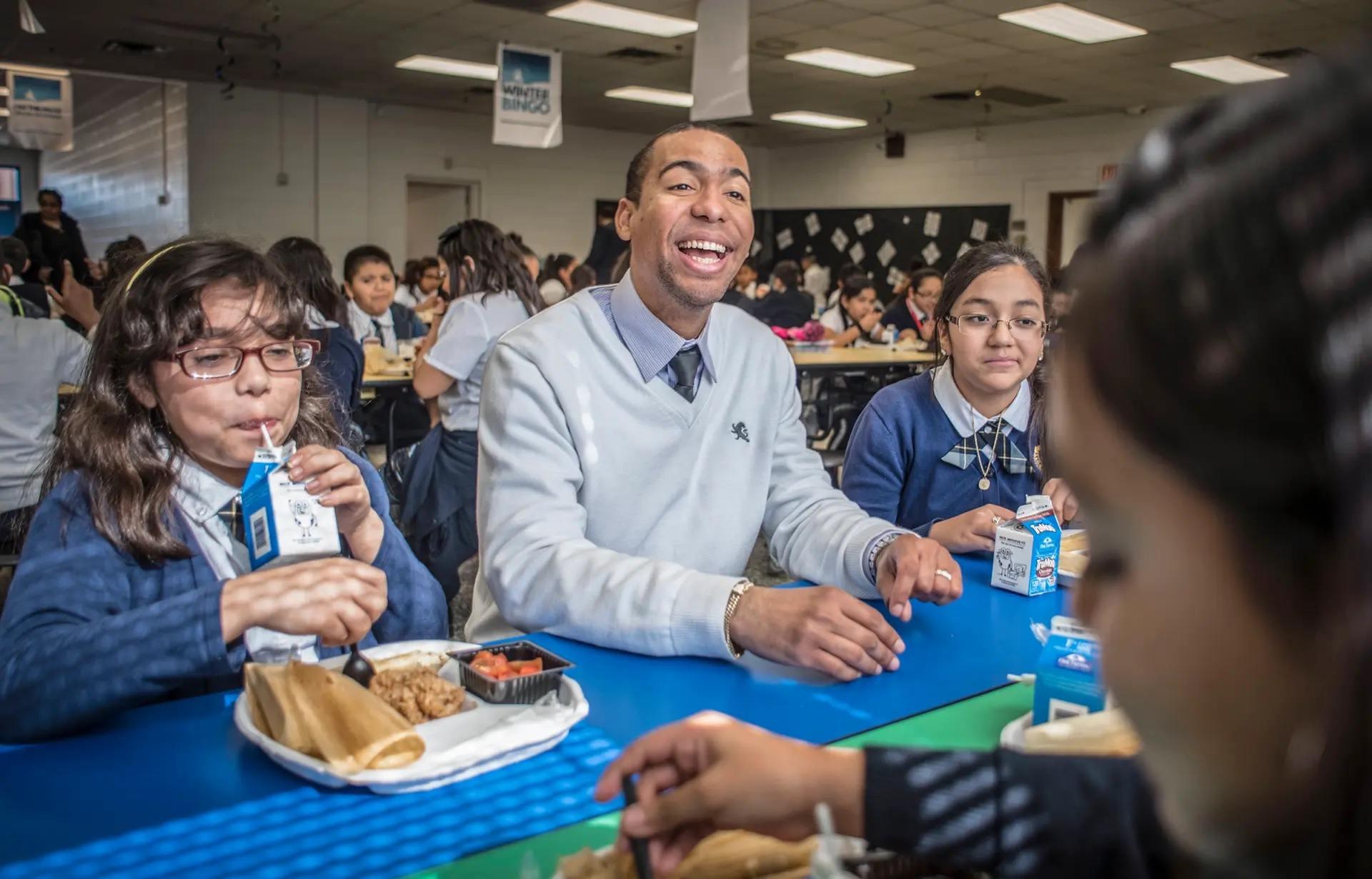An elementary teacher sits and laughs with students at the lunch table