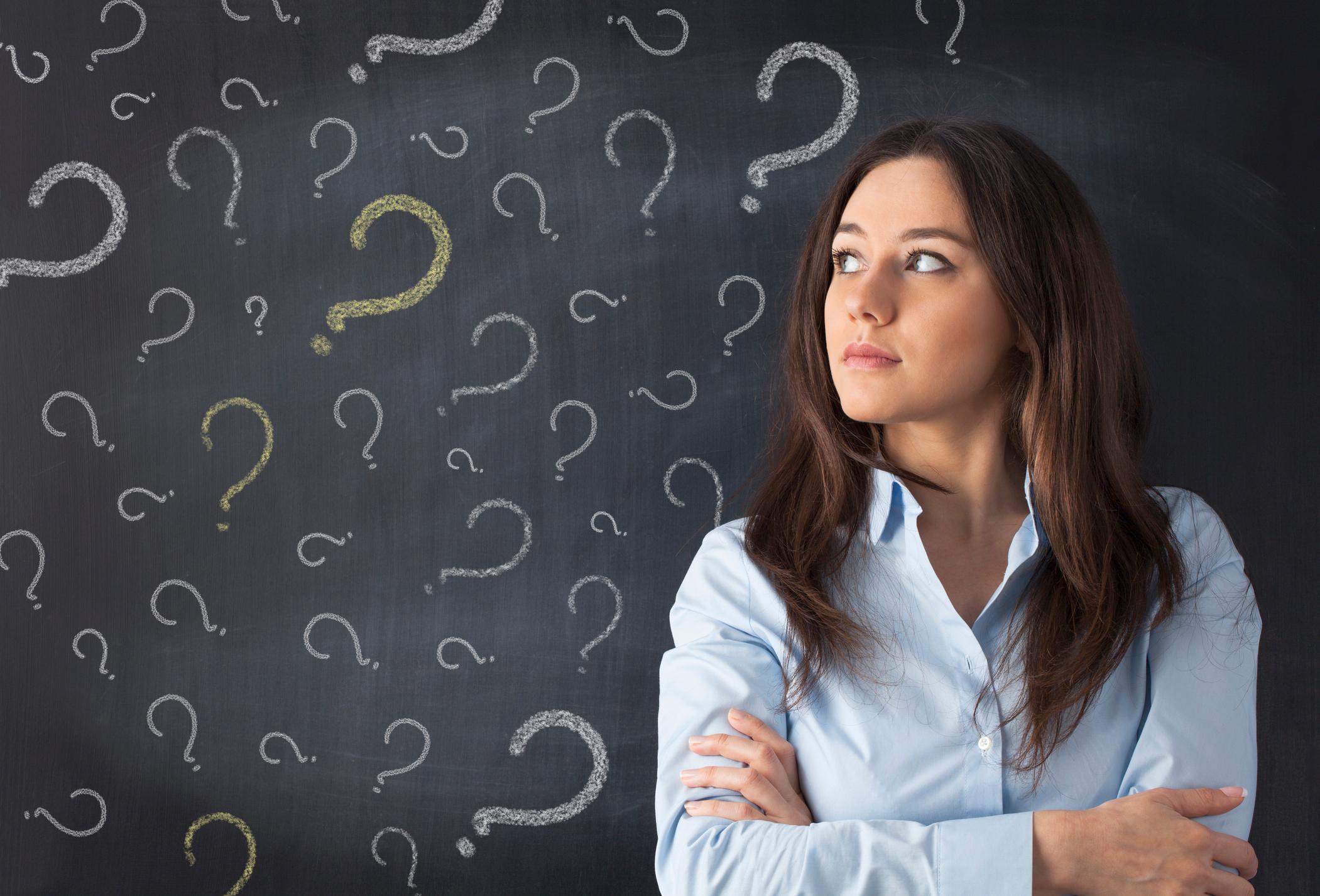 Female college student with arms crossed looking pensive, in front of chalk board with question marks on it